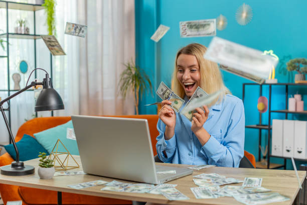 Money cash dollar rain falling on Caucasian young woman using laptop celebrating success play winning game lottery jackpot casino online goal achievement good news luck. Girl sits at home office table